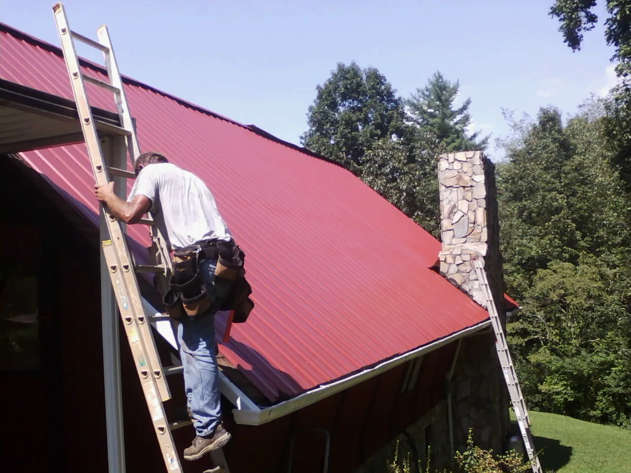 Roofer on a ladder beside a red metal roof and stone chimney — Soffit & Fascia Repair by Roofing Brothers in Yellow Pine